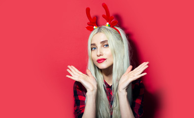 Portrait of young blonde girl with red lips and blue eyes, wearing a Christmas deer horns and plaid shirt, isolated on red background.
