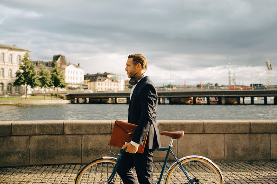 Side view of businessman walking with bicycle on footpath in city against sky