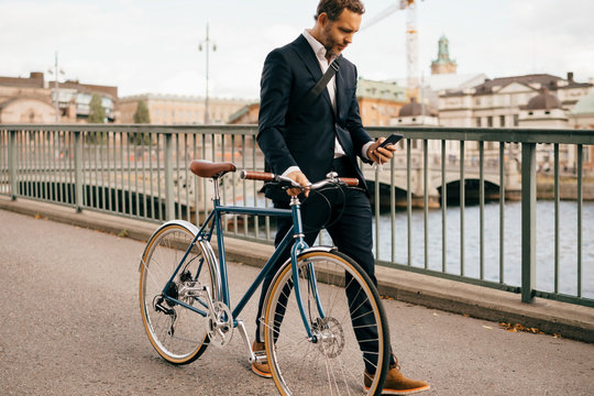 Confident businessman using smart phone while walking with bicycle on bridge in city - Powered by Adobe