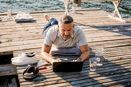 Mature man lying on front while using laptop over wooden jetty