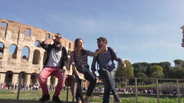 Three young friends tourists sitting in front of colosseum in rome talking having fun near arch of constantine with backpacks sunglasses happy beautiful girl long hair