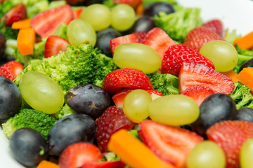 Closeup of fruit salad with broccoli, green and black grapes, strawberry slices and sliced ​​carrots and cubes. Delicious, nutritious and healthy meal in white ceramic serving dish on the wooden table
