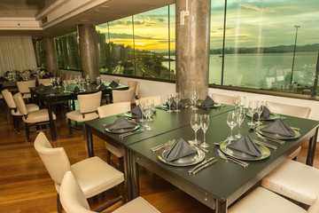 Luxurious party room with various tables, plates, cutlery and wine glasses. Windows overlooking the sea and sunset. Hall for events, parties, celebrations with beautiful view.
