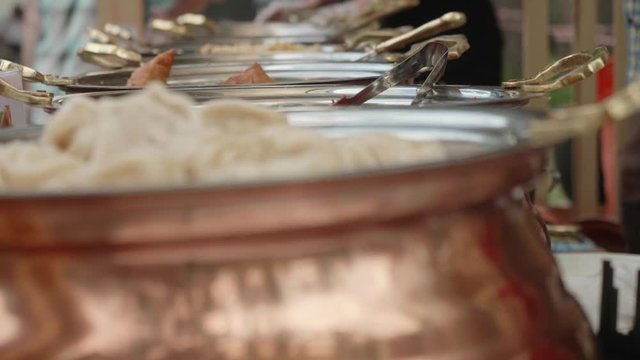 The row of casseroles with pelmeni, rice and other asian food. Background is in focus. Hands of cooks cooking food.