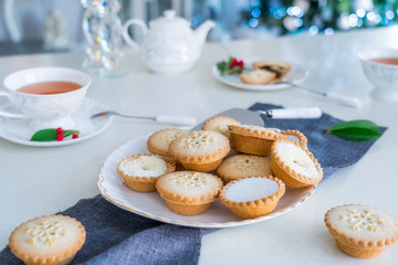 Traditional english festive pastry mince pies on served for tea time table with lightened christmas tree on background. Cozy home mood. Close up, selective focus. Copy space.