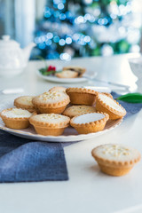 Obraz na płótnie Canvas Traditional english festive pastry mince pies on served for tea time table with lightened christmas tree on background. Cozy home mood. Vertical card. Close up, selective focus. Copy space.