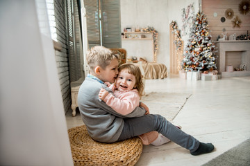 Brother and sister in the bright living room at home on the bed. small children friends in hats, wrapped in a warm blanket, playing with a Teddy bear on the background of Christmas decor.