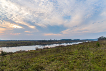 The mirror water reflects trees and the colorful glow of sunset in a cloudy sky. Beautiful spacious landscape made at dusk from the high bank of a river. Natural background