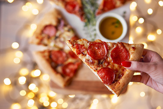 Christmas and New Year atmosphere.Womans hand takes slice of Italian pizza with melting tomato, pepperoni and cheese on a white marbel cutting board.Background with lights in bokeh and selective focus