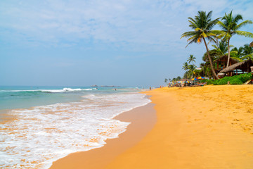 Hikkaduwa, Sri Lanka. March 8, 2018. Beach on the Indian Ocean. Sunny day, yellow sand, palm trees and foam waves.
