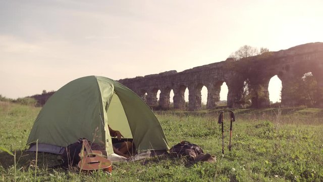 Young man backpacker wakes up exits camping tent in front of roman aqueduct arches in parco degli acquedotti park ruins in rome at sunset sticks in ground slow motion