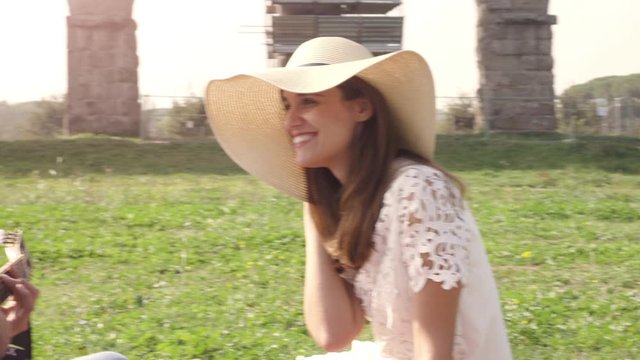 Happy young lovely couple sitting lying on a blanket in the grass in front of ancient roman aqueduct ruins in parco degli acquedotti park in rome romantic play guitar sing beautiful girl with hat