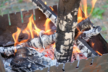 Metal portable barbecue on the ground in the yard. Brazier is covered with rust. Firewood burns out in the barbecue, a bright orange fire. One log in the barbecue, outdoor recreation barbecue grill.