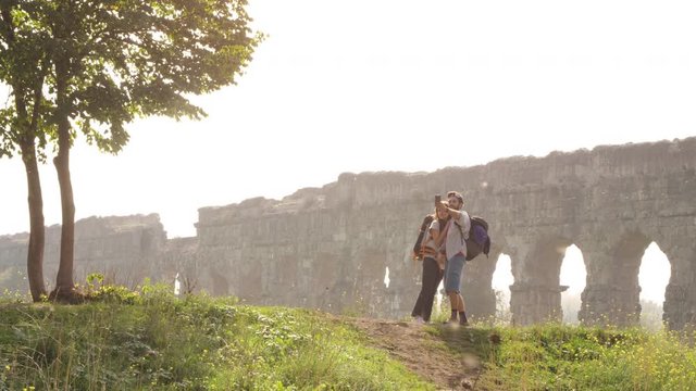 Young lovely couple backpackers tourists taking selfies photo on hill in front of roman aqueduct arches in parco degli acquedotti park ruins in rome on romantic misty sunrise with sleeping bag slow