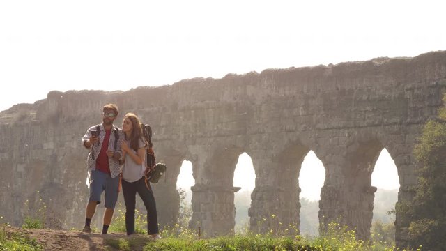 Young lovely couple backpackers tourists taking selfies photo on hill in front of roman aqueduct arches in parco degli acquedotti park ruins in rome on romantic misty sunrise with sleeping bag slow