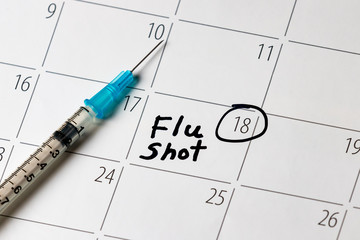 Flu shot reminder written in black in on white calendar. Syringe with needle in background. Influenza prevention and healthcare concept