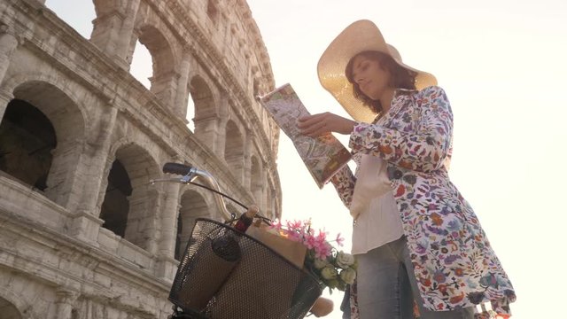 Beautiful young woman in colorful fashion dress with bike reading map in front of colosseum in Rome at sunset with happy attractive tourist girl with straw hat looking for directions ground shot dolly