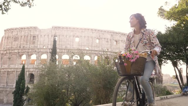 Beautiful young woman in colorful fashion riding bike in front of colosseum in Rome at sunset with trees happy attractive girl tourist with straw hat in colle oppio front view steadycam dolly