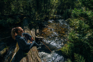 Beautiful girl sits on log and chilling after travel. Woman traveler in summer passes on wooden bridge in background of forest. Bridged lies on the river. Model of caucasian is chill and happy.