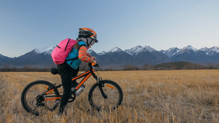Obraz premium One caucasian children walk with bike in wheat field. Little girl walking black orange cycle on background of beautiful snowy mountains. Biker stand with backpack and helmet. Mountain bike hardtail.