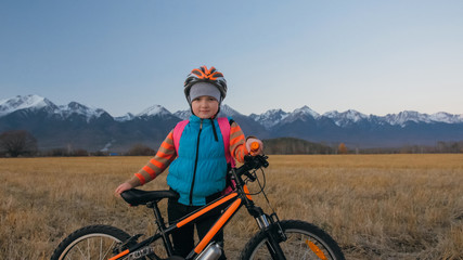 One caucasian children walk with bike in wheat field. Little girl walking black orange cycle on background of beautiful snowy mountains. Biker stand with backpack and helmet. Mountain bike hardtail.