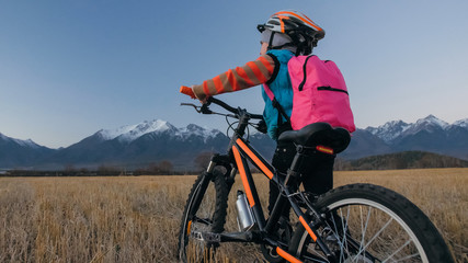 Obraz premium One caucasian children walk with bike in wheat field. Little girl walking black orange cycle on background of beautiful snowy mountains. Biker stand with backpack and helmet. Mountain bike hardtail.