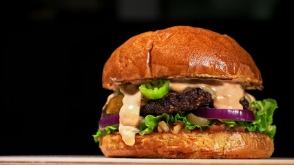 Craft burger is cooking on black background. Consist: red sauce salsa, lettuce, red onion, pickle, cheese, chilli green pepper, air bun and marbled meat beef. Not made ideal. Loving hand made.