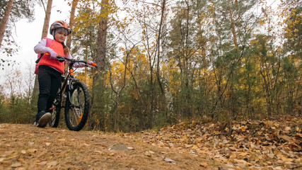 Naklejka premium One caucasian children walk with bike in autumn park. Little girl walking black orange cycle in forest. Kid goes do bicycle sports. Biker motion ride with backpack and helmet. Mountain bike hardtail.