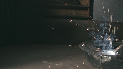 Forge workshop. Smithy. Worker in a welding hood helmet welds a part by electric welding. Sparks are reflected in the protective screen. Blacksmith makes iron product for manufacture. Slow motion.