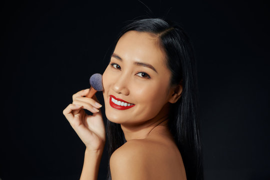 Beauty portrait of smiling half-naked woman with fresh skin applying makeup with soft brush isolated over black background