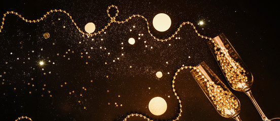 Champagne glasses filled with golden decorations on a black shiny background. Celebration minimal...