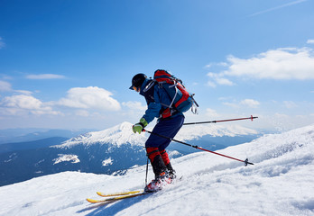 Fototapeta na wymiar Sportsman skier in bright clothing, helmet and goggles with backpack riding down steep snowy slope on copy space background of blue sky and mountain landscape. Winter holidays, extreme sport concept.