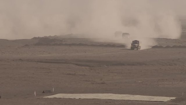 Military cargo convoy rides fast in desert sand dunes, leaving behind a massive trail of dust, military operation. The convoy group is moving in formation to meet the combat attack
