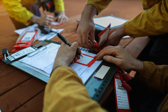 Construction miners worker writing his name and sign onto personnel red danger tag and placing locking into isolation permit safety control lockbox while his friends locking on at the background