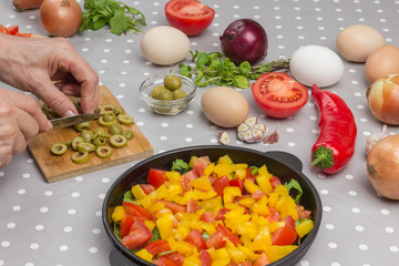 Sliced tomatoes and yellow pepper in a pan. Hands chopped olives. Eggs, red pepper, onions, olives, garlic, mint on the table.
