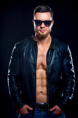 Fototapeta premium Young handsome macho man in sunglasses with open leather jacket revealing muscular chest and torso, looking to the camera. closeup