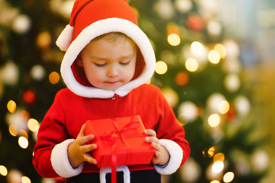 Happy child holding red gift box on the background of Christmas lights. Smiling funny kid in santa costume holds christmas present in hands. Christmas and New year concept. Christmas children.