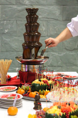 Chocolate fondue fountain with cook hand. Food photography with fruit snacks. Catering service at the celebration event. Melted sweet chocolate. Gray stone background