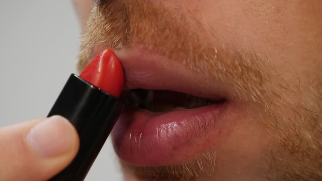 Unrecognized gay man is painting lips with red lipstick, close up side view. Travesty putting red lipstick on face. Guy applying transsexual makeup