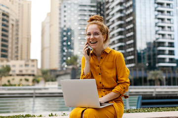 Young Woman sitting in Dubai Marina aria, talking on the phone and work on laptop. Student or ...