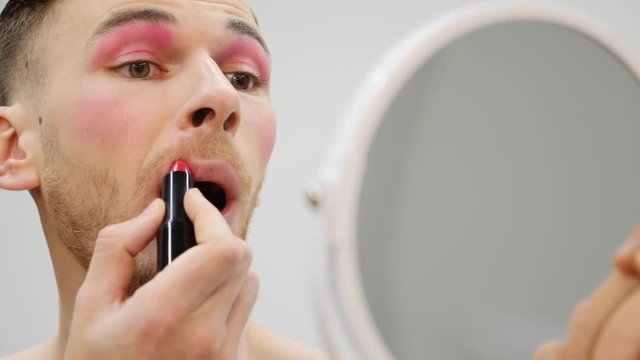 Travesty man applies lipstick on lips. Transsexual guy painting lips with pink lipstick. Young man making travesty makeup, close up