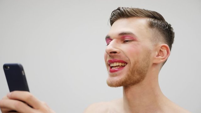Young travesty man is texting on smartphone and smiling, close up side view. Pretty transsexual making selfies on phone. Young gay man with pink makeup looking at phone