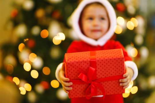 Christmas gift in the hands of a child. Hands of little boy in santa costume with Christmas present. Smiling funny kid holding red gift box on the background of lights. Holidays, New year concept.