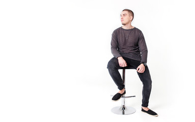 A young attractive guy in gray and black clothes sits on a bar stool on a white background and...
