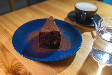 Snickers cheesecake on a blue saucer, a glass of water and a cup of fresh cappuccino on a large bright wooden table