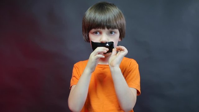 A child in an orange T-shirt peels off a black adhesive tape from his face.
