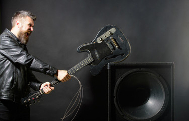 Angry man with beard breaks guitar at music speakers. Emotions, aggression, musical instruments. Guitar player at rock concert. Musician with electric guitar. Guitarist breaks guitar at music column.