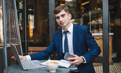 Young businessman with smartphone sitting at desk with laptop and looking at camera