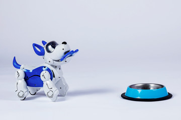 Electronic interactive toy dog puppy with a bone in mouth stands in front of empty pet bowl. High technology concept of future domestic animals in electronic home. Indoors, copy space.