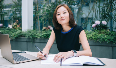 Cheerful young beautiful freelance woman sitting writing notes while working on laptop in workplace